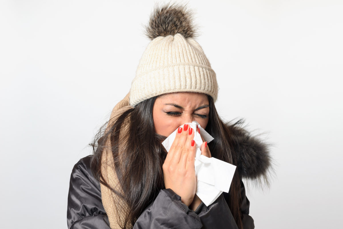 Young woman with a seasonal winter cold and flu dressed in warm trendy clothing blowing her nose on a white handkerchief
